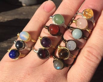 May include: A collection of 12 rings featuring different colored gemstones wrapped in gold wire. The gemstones include amethyst, carnelian, aquamarine, labradorite, tiger's eye, moonstone, lapis lazuli, hematite, rose quartz, citrine, and aventurine.