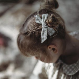 May include: A young girl with blonde hair wearing a white dress with a gingham pattern and a white bow in her hair.