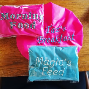 Op de afbeelding: Drie kleurrijke zakjes met geborduurde tekst. Een felroze zakje heeft "Morning Food" in blauw-wit gestreepte letters. Een ander roze zakje heeft "Taz's Breakfast" in blauw script. Een blauw zakje leest "Magic's Feed" in wit script.