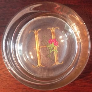 May include: A clear glass coaster with a gold embroidered letter H and a red flower design.
