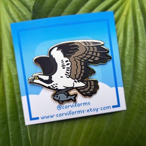 May include: A gold-colored enamel pin featuring a cartoon osprey with brown and black wings, white body, and yellow talons, holding a fish in its beak. The pin is on a blue background with white clouds and the text "@corviforms www.corviforms.etsy.com".
