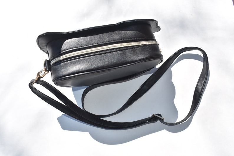 May include: Black faux leather shoulder bag with a rounded shape and a zipper closure. The bag has a long adjustable strap.