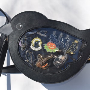 May include: Black faux leather crow-shaped crossbody bag with a clear plastic window displaying a variety of crow-themed enamel pins. The pins include a crow with a crown, a crow with a pumpkin, and a crow with the words "Never Crow" written on a scroll.