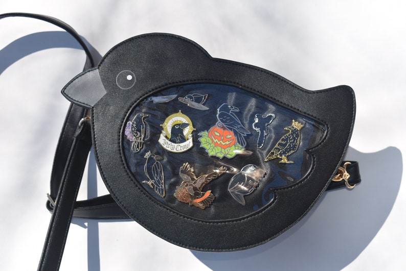May include: Black faux leather crow-shaped crossbody bag with a clear plastic window displaying a variety of crow-themed enamel pins. The pins include a crow with a crown, a crow with a pumpkin, and a crow with the words "Never Crow" written on a scroll.