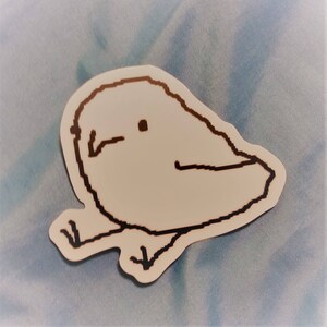 以下が含まれることがあります： 黒いアウトラインで描かれた、シンプルな漫画風の鳥のイラストが特徴の白いステッカー。鳥は小さな嘴、1つの目、2本の足を持っています。ステッカーは水色の布の背景にあります。