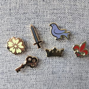 Op de afbeelding: Set van zeven gouden emaille pinnen met een bloem, een zwaard, een duif, een kroon, een sleutel en een lelie.