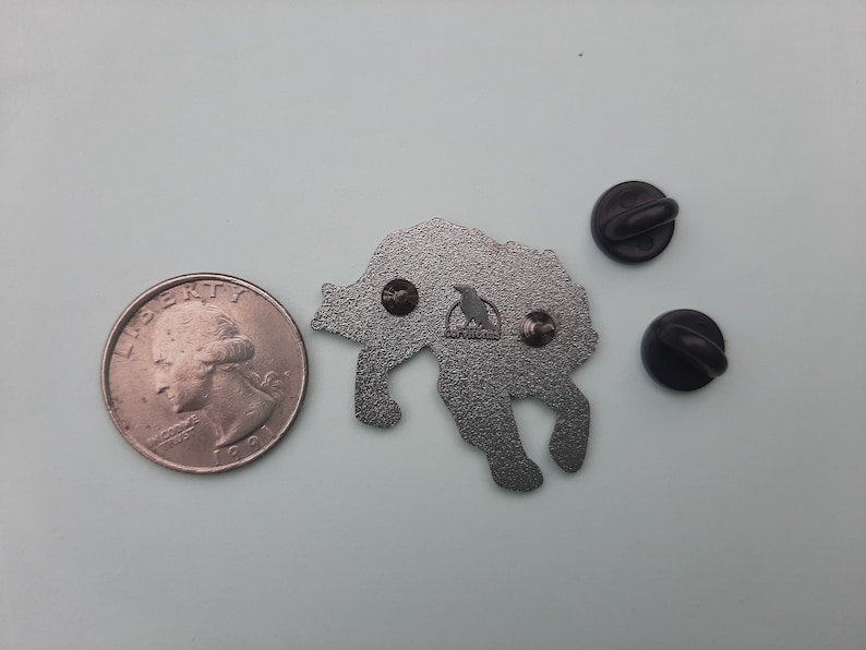 K&ouml;nnte beinhalten: Eine silberne Metallstifte mit einem Wolf, der zum Mond heult. Die Stifte ist etwa so gro&szlig; wie eine 25-Cent-M&uuml;nze. Die R&uuml;ckseite der Stifte hat ein schwarzes Vogellogo und zwei Metallstecker.