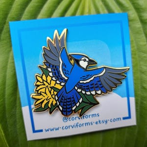 Può includere: Spilla smaltata a forma di ghiandaia azzurra con dettagli dorati, in volo con le ali spiegate, posata su un fiore giallo con foglie verdi. La spilla è su uno sfondo blu con il testo "@corviforms www.corviforms.etsy.com".