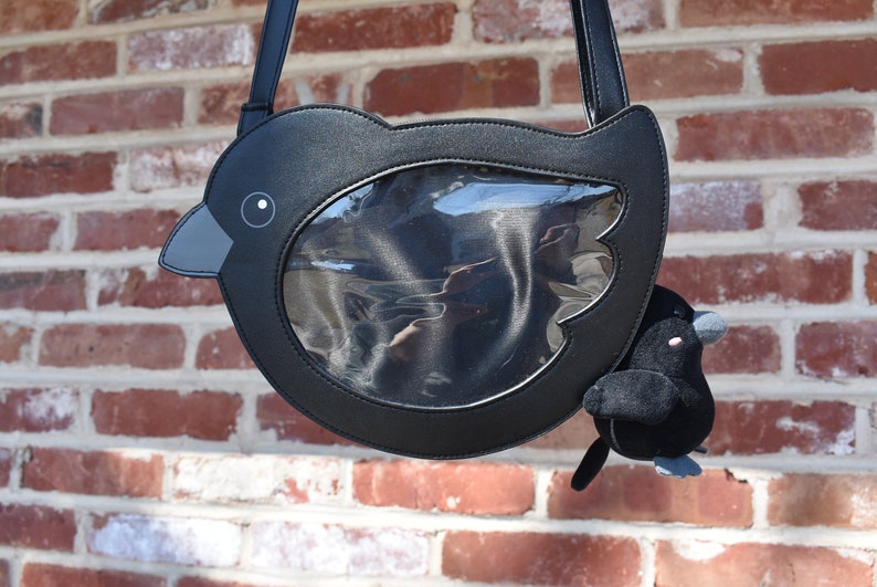 May include: A black faux leather bird-shaped crossbody bag with a clear plastic window on the front. A small black plush bird is attached to the bag.