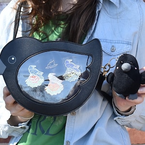 May include: A black faux leather bird-shaped purse with a clear plastic window showing a variety of colorful pins. The purse has a black plush bird keychain attached.