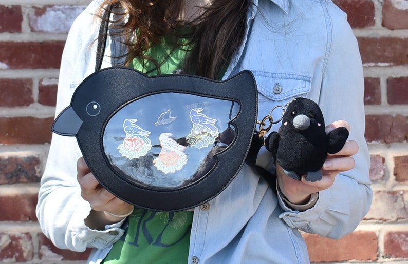 May include: A black faux leather bird-shaped purse with a clear plastic window showing a variety of colorful pins. The purse has a black plush bird keychain attached.