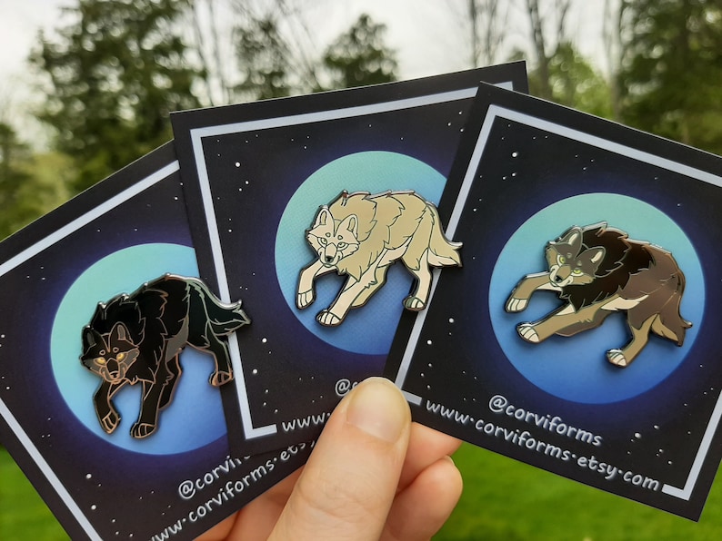 K&ouml;nnte beinhalten: Drei Emaille-Pins mit verschiedenen Wolfsdesigns. Jeder Pin zeigt einen Wolf, der zum Mond heult, vor einem Hintergrund eines sternenklaren Nachthimmels. Die Wolfsdesigns sind schwarz, wei&szlig; und braun.