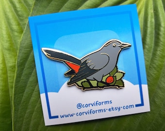 May include: A gray and black enamel pin of a gray catbird perched on a branch with a red berry. The pin has a gold outline and is on a blue background with white clouds. The text "@corviforms" and "www.corviforms.etsy.com" is at the bottom of the pin.