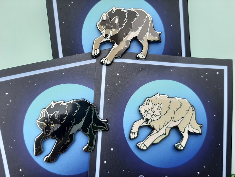 K&ouml;nnte beinhalten: Drei Emaille-Pins mit W&ouml;lfen, die zum Mond heulen. Die W&ouml;lfe sind in verschiedenen Farben dargestellt: Schwarz, Grau und Wei&szlig;. Der Mond ist blau mit einem sternen&uuml;bers&auml;ten Hintergrund.