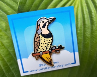 May include: A colorful enamel pin featuring a stylized woodpecker perched on a branch. The bird has a black, tan, and yellow body with a gray head and a black beak. The pin is set against a blue background.