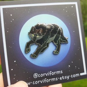 K&ouml;nnte beinhalten: Schwarze Emaille-Anstecknadel mit einem Wolf mit gelben Augen, der auf einem blauen und wei&szlig;en Mondhintergrund steht. Die Anstecknadel befindet sich auf einer schwarzen Karte mit wei&szlig;en Sternen und dem Text "@corviforms w-corviforms.etsy.com".