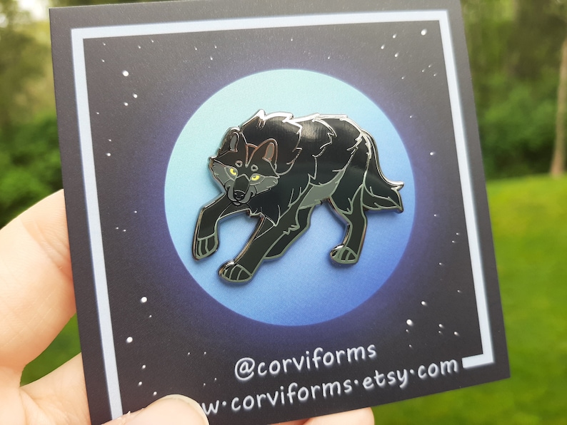 K&ouml;nnte beinhalten: Schwarze Emaille-Anstecknadel mit einem Wolf mit gelben Augen, der auf einem blauen und wei&szlig;en Mondhintergrund steht. Die Anstecknadel befindet sich auf einer schwarzen Karte mit wei&szlig;en Sternen und dem Text "@corviforms w-corviforms.etsy.com".