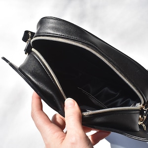 May include: Black leather crossbody bag with a zipper closure and a gold-toned zipper pull. The bag is open and the interior is lined with black fabric.