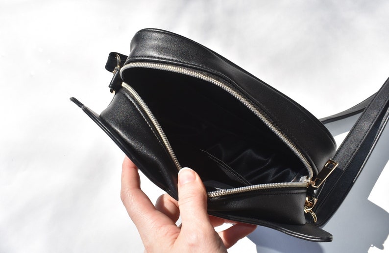 May include: Black leather crossbody bag with a zipper closure and a gold-toned zipper pull. The bag is open and the interior is lined with black fabric.