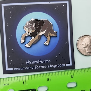 K&ouml;nnte beinhalten: Eine braune und graue Wolf-Emaille-Anstecknadel mit gelben Augen, vor einem Hintergrund aus blauem Mond mit wei&szlig;en Sternen. Die Anstecknadel hat einen Durchmesser von etwa 2,5 cm. Die Anstecknadel befindet sich auf einer wei&szlig;en Karte mit dem Text "@corviforms www.corviforms.etsy.com".