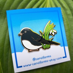 May include: Gold enamel pin featuring a black and white bird with a white breast and a black head and back. The bird is perched on a green stem with white flowers. The pin has a blue background and the text "@corviforms www.corviforms.etsy.com" at the bottom.
