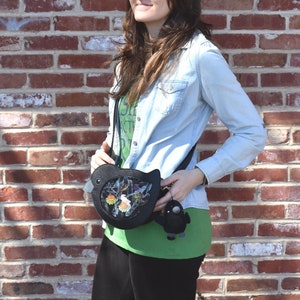 May include: A black bird-shaped crossbody bag with a clear window showing colorful details. The bag is worn by a person wearing a blue denim shirt and a green shirt with text.