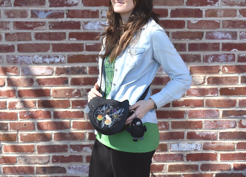 May include: A black bird-shaped crossbody bag with a clear window showing colorful details. The bag is worn by a person wearing a blue denim shirt and a green shirt with text.