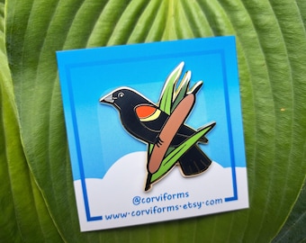 Op de afbeelding: Een zwarte en gouden emaille pin met een roodvleugelzwarte vogel op lisdodden. De pin staat op een blauwe kaart met de naam en website van de winkel. De achtergrond is een groot groen blad.