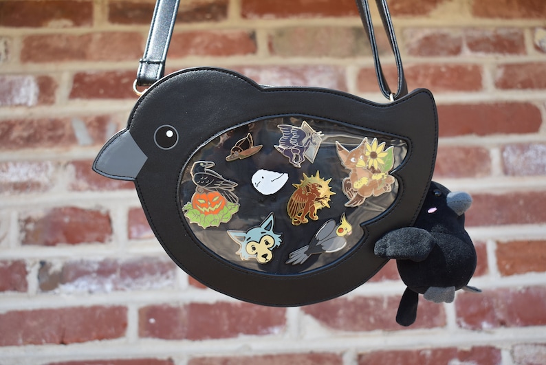 May include: A black crow-shaped purse with a clear plastic window displaying various enamel pins. The pins feature a variety of designs, including a crow, a ghost, a pumpkin, a sunflower, and a bird. A small black plush crow is attached to the purse.
