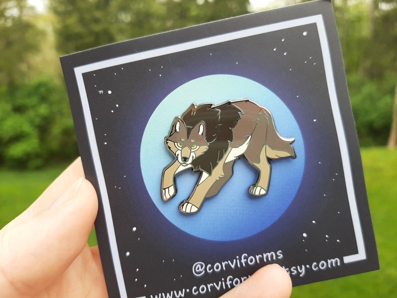 K&ouml;nnte beinhalten: Eine silberne Emaille-Anstecknadel mit einem braunen Wolf mit gelben Augen, der auf einem blauen Mondhintergrund mit wei&szlig;en Sternen steht. Die Anstecknadel befindet sich auf einer schwarzen Karte mit dem Text "@corviforms www.corviforms.etsy.com".