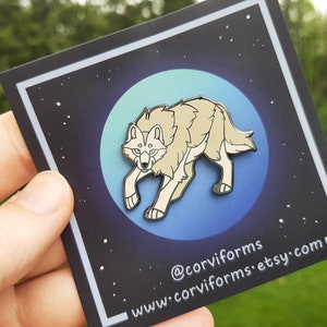 K&ouml;nnte beinhalten: Eine silberne Emaille-Anstecknadel mit einem wei&szlig;en Wolf vor einem blauen und gr&uuml;nen Mondhintergrund. Die Nadel hat einen schwarzen Rand und der Text "@corviforms www.corviforms.etsy.com" ist unten aufgedruckt.