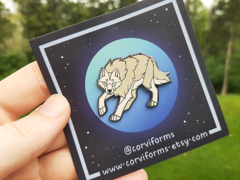 K&ouml;nnte beinhalten: Eine silberne Emaille-Anstecknadel mit einem wei&szlig;en Wolf vor einem blauen und gr&uuml;nen Mondhintergrund. Die Nadel hat einen schwarzen Rand und der Text "@corviforms www.corviforms.etsy.com" ist unten aufgedruckt.