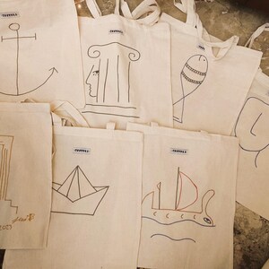 May include: A selection of off-white tote bags, each featuring a unique hand-drawn design in black and brown ink. Designs include an anchor, a face with columns, a fish, a simple face, a building, a paper boat, and a sailboat. Each bag has a small label.