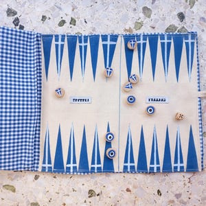 Op de afbeelding: Een opgerold backgammonbord met een blauw en wit geruit exterieur. Het speeloppervlak is crèmekleurig met blauwe driehoeken en het woord "SETTLE" erop gedrukt. Houten speelstukken en een dobbelsteen liggen op het bord.
