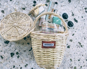 Könnte beinhalten: Ein natürlicher Weidenkorb mit Henkel, gefüllt mit Schönheitsprodukten und Accessoires. Der Korb hat ein rechteckiges Etikett mit dem Text "SATTALI". Ein runder Weidenkorbdeckel liegt neben dem Korb. Weitere Artikel sind Sonnenbrillen, Make-up-Pinsel und eine kleine Kosmetiktasche.
