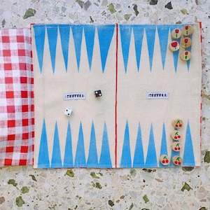May include: A backgammon board with a red and white checkered fabric on the left side. The board has a cream-colored background with blue triangles and red lines. Dice and wooden game pieces with flower and cherry designs are scattered on the board.