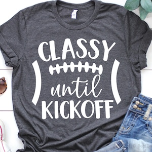 May include: Dark gray t-shirt with the words "CLASSY until KICKOFF" in white, designed in the shape of a football. The shirt is made of a soft material and is suitable for casual wear. The shirt is folded, with sunglasses and denim shorts visible.