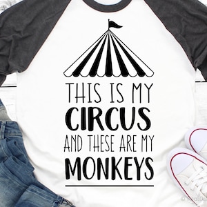 Op de afbeelding: Een wit en grijs raglan t-shirt met een zwart-witte afbeelding van een circus tent en de tekst "This is my circus and these are my monkeys".