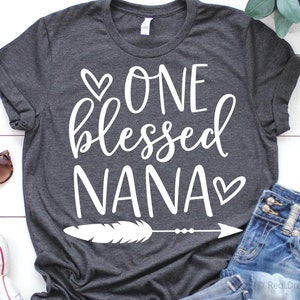 May include: A dark grey t-shirt with the text "ONE blessed NANA" in white lettering with a small heart on either side of the text. A white feather with an arrow pointing to the right is below the text.