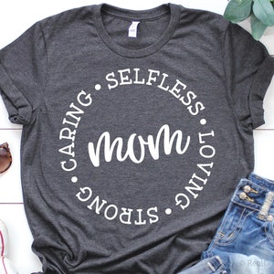 May include: A dark gray t-shirt with a white circle graphic that says "mom" in the center and the words "caring", "strong", "loving", and "selfless" around the circle.