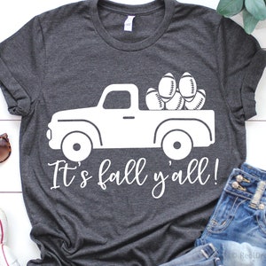 May include: A dark gray t-shirt with a white graphic of a pickup truck carrying footballs. The text "It's fall y'all!" is printed below the truck.