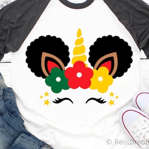 May include: A white t-shirt with a black and brown unicorn design. The unicorn has a gold horn and is wearing a crown of red, yellow, and green flowers. There are gold stars around the unicorn's face.