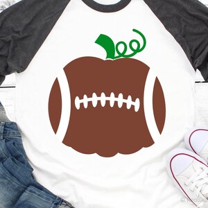Op de afbeelding: Een wit T-shirt met een bruine en witte voetbal in de vorm van een pompoen met een groene steel en blad. Het shirt is raglan met een grijze mouw.