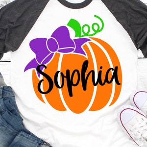 May include: A white t-shirt with an orange pumpkin with a purple bow and green stem. The name "Sophia" is written in black cursive on the pumpkin.