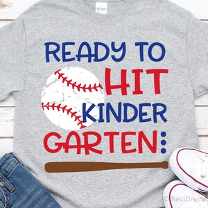 May include: Grey t-shirt with the text "Ready to Hit Kinder Garten:" in red and blue lettering. A baseball with red stitching is next to the text. A brown baseball bat is below the text.