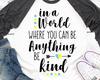May include: A white and grey baseball tee shirt with the text "in a world where you can be anything be kind" in black lettering. The text is surrounded by yellow and blue dots.