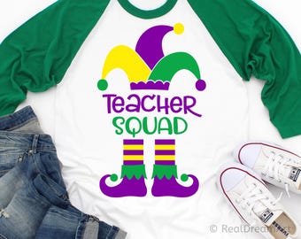 May include: White raglan-style t-shirt with green sleeves. The shirt features a colorful graphic of a jester's hat and elf shoes, with the words "Teacher Squad" in purple and green. A pair of white sneakers and blue jeans are also visible.