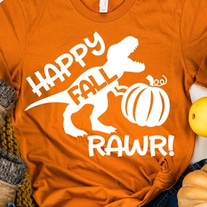 Puede incluir: Camiseta naranja con un gráfico de dinosaurio blanco sosteniendo una calabaza y el texto "Happy Fall Rawr!"