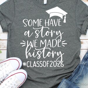 Pu&ograve; includere: Maglietta grigia con testo bianco e grafica del cappello di laurea. Il testo recita "SOME HAVE a STORY WE MADE history #CLASSOF2026". Sono visibili anche scarpe da ginnastica bianche e jeans blu.