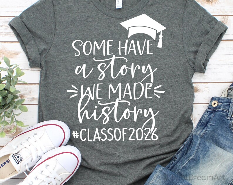 Pu&ograve; includere: Maglietta grigia con testo bianco e grafica del cappello di laurea. Il testo recita "SOME HAVE a STORY WE MADE history #CLASSOF2026". Sono visibili anche scarpe da ginnastica bianche e jeans blu.
